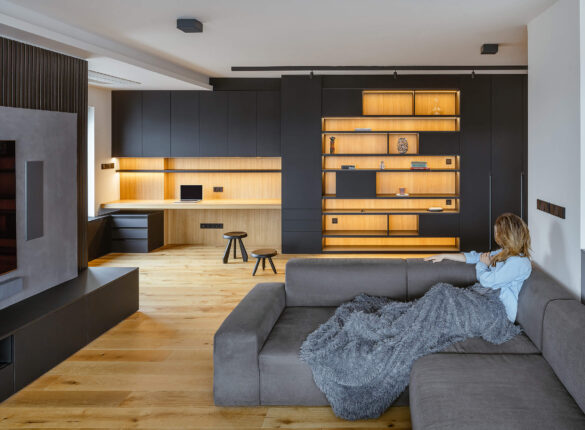 Propojený prostor kuchyně a obývacího pokoje s minimalistickým designem, dřevěnou podlahou, šedými a černými akcenty a integrovaným osvětlením.