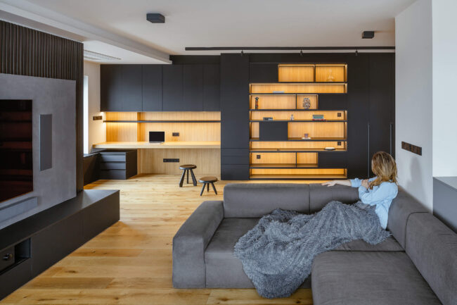 Propojený prostor kuchyně a obývacího pokoje s minimalistickým designem, dřevěnou podlahou, šedými a černými akcenty a integrovaným osvětlením.