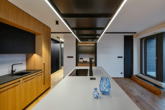 Propojený prostor kuchyně a obývacího pokoje s minimalistickým designem, dřevěnou podlahou, šedými a černými akcenty a integrovaným osvětlením.